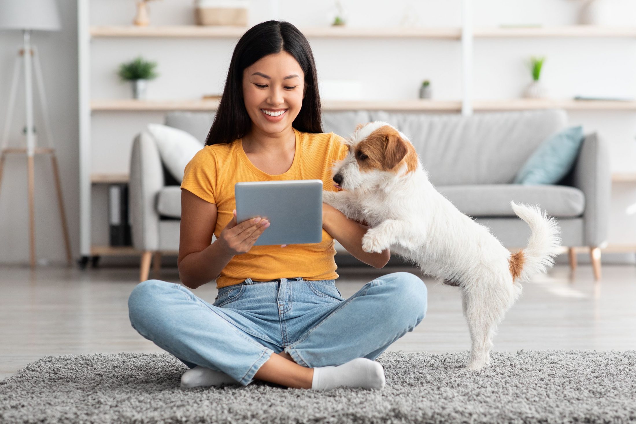 A dog leans on his owner as she sits on the floor with her tablet.